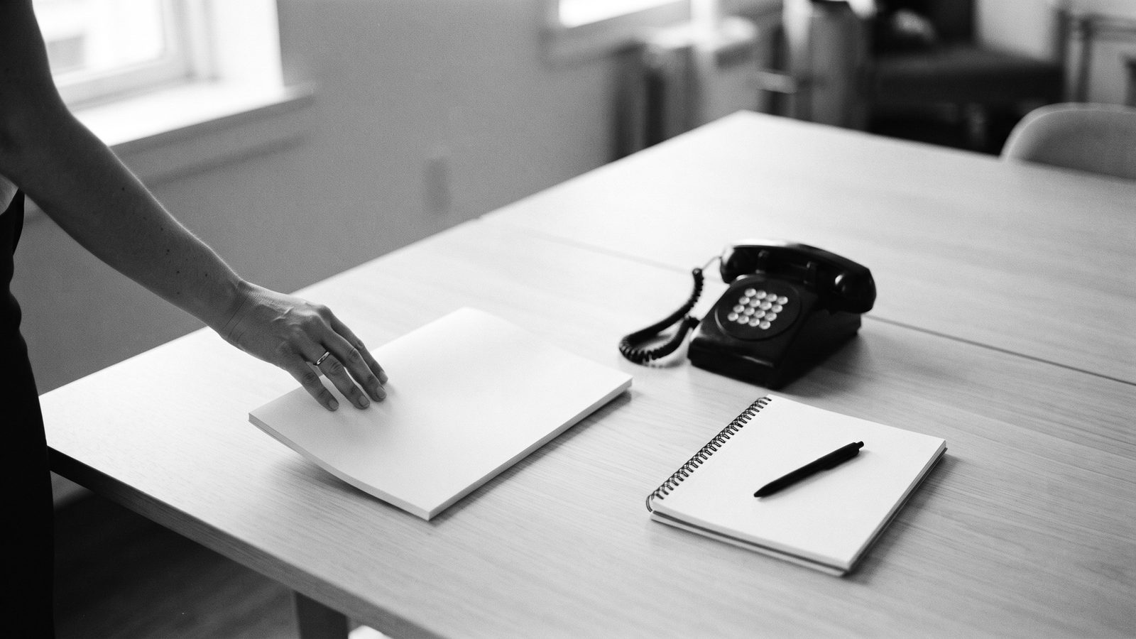 Eine Hand legt Unterlagen auf einen leeren hellen Tisch, daneben Telefon und Notizblock