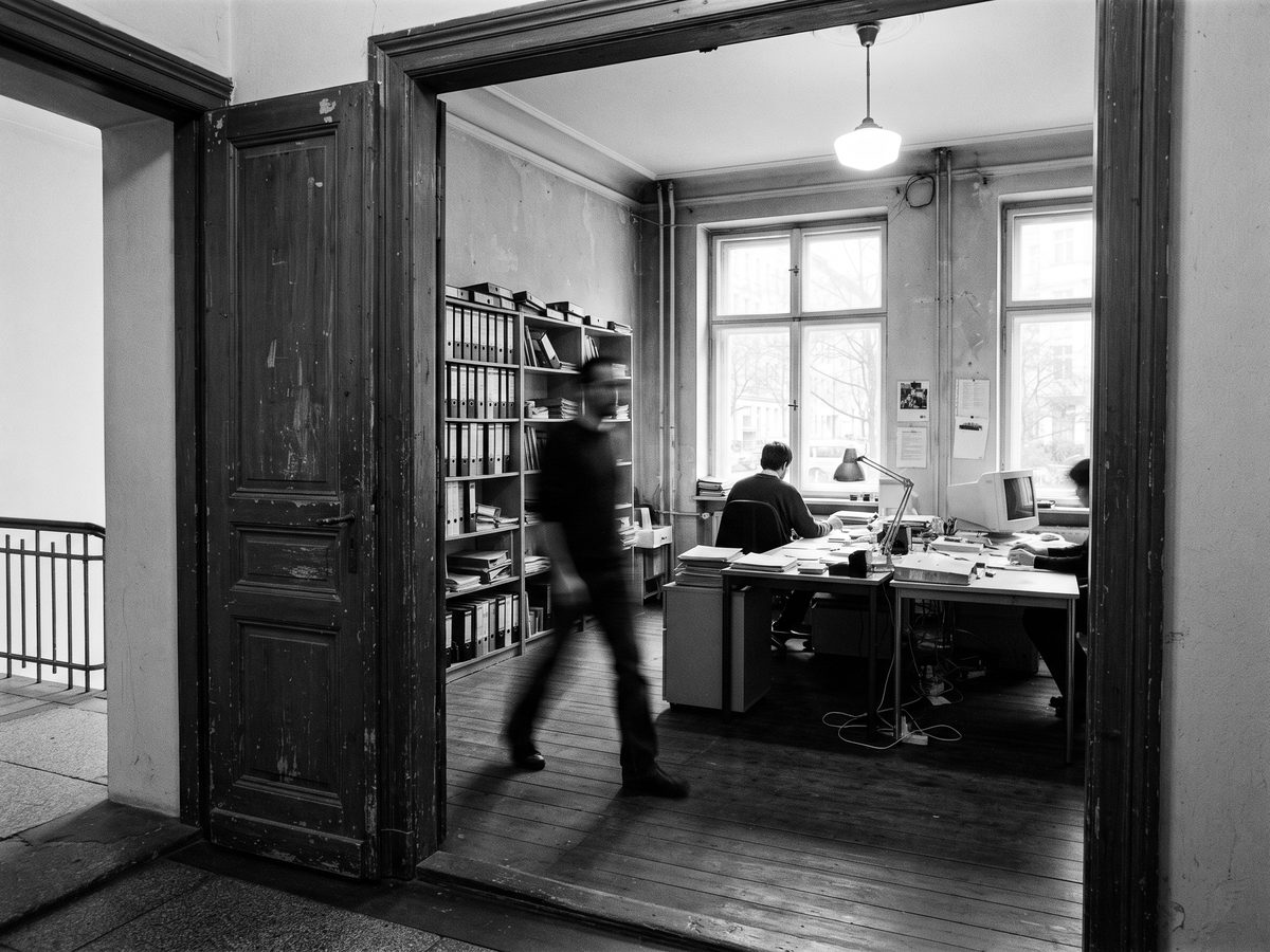 Blick durch eine offene Altbautür in ein Büro mit hohen Fenstern und Aktenregal