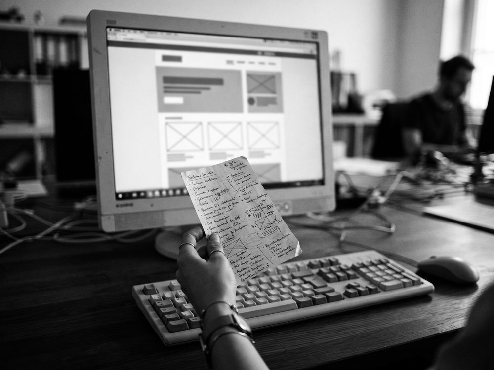 Bildschirm mit einem angedeuteten Seitenlayout, davor eine Hand mit Notizzettel